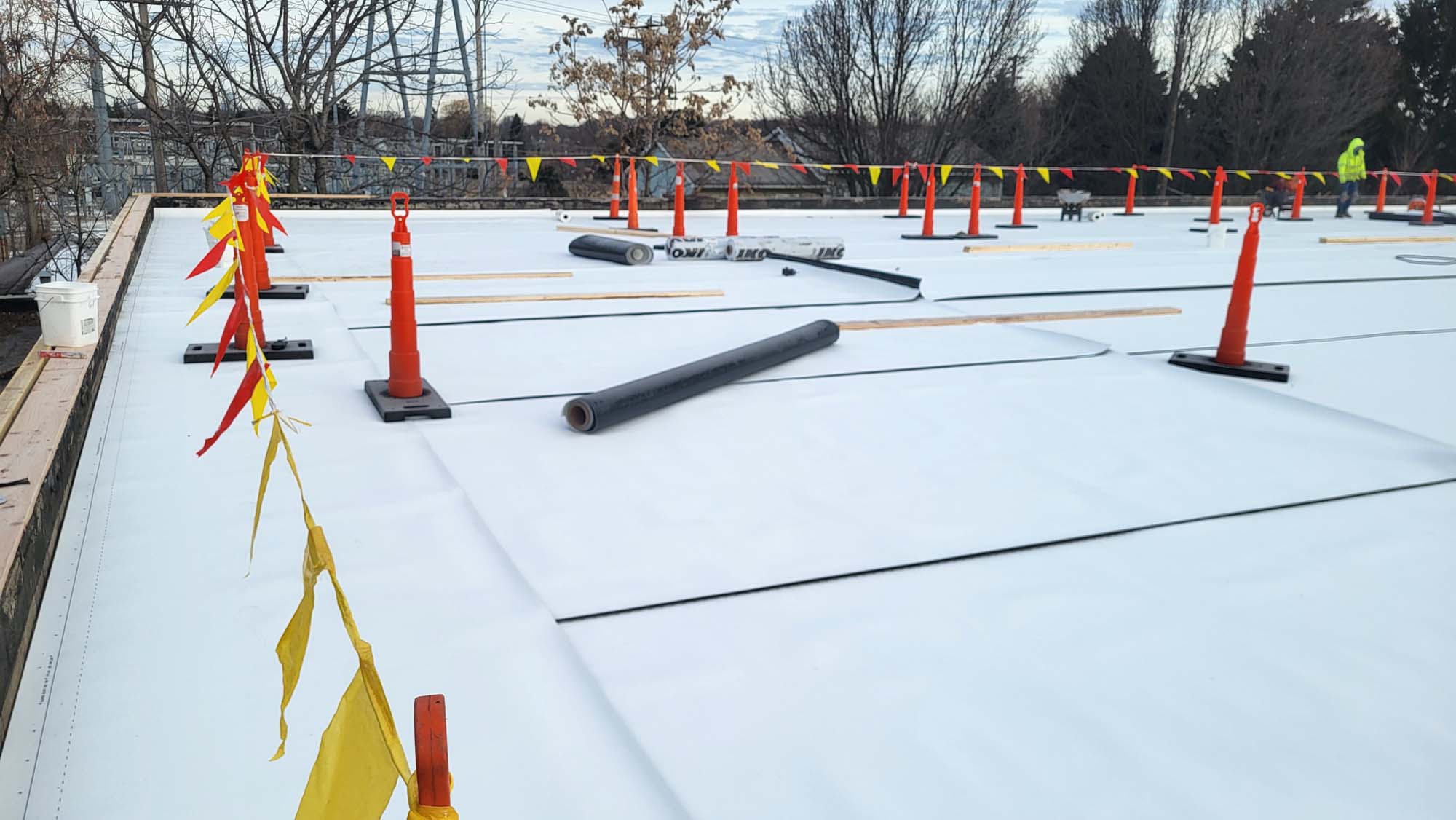 A flat rooftop under construction with a white surface, orange safety cones, yellow caution tape, some pipes, and trees in the background. The sky is partly cloudy.
