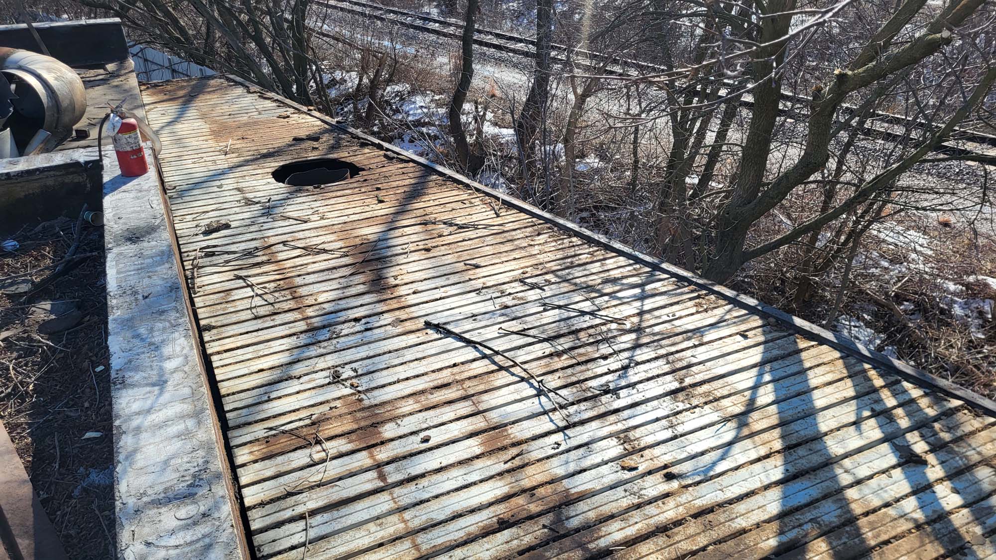 A rusted, weathered metal surface with a hole sits outdoors, surrounded by leafless trees and bushes. A red bottle stands on the left, and train tracks run in the background amid patches of snow.