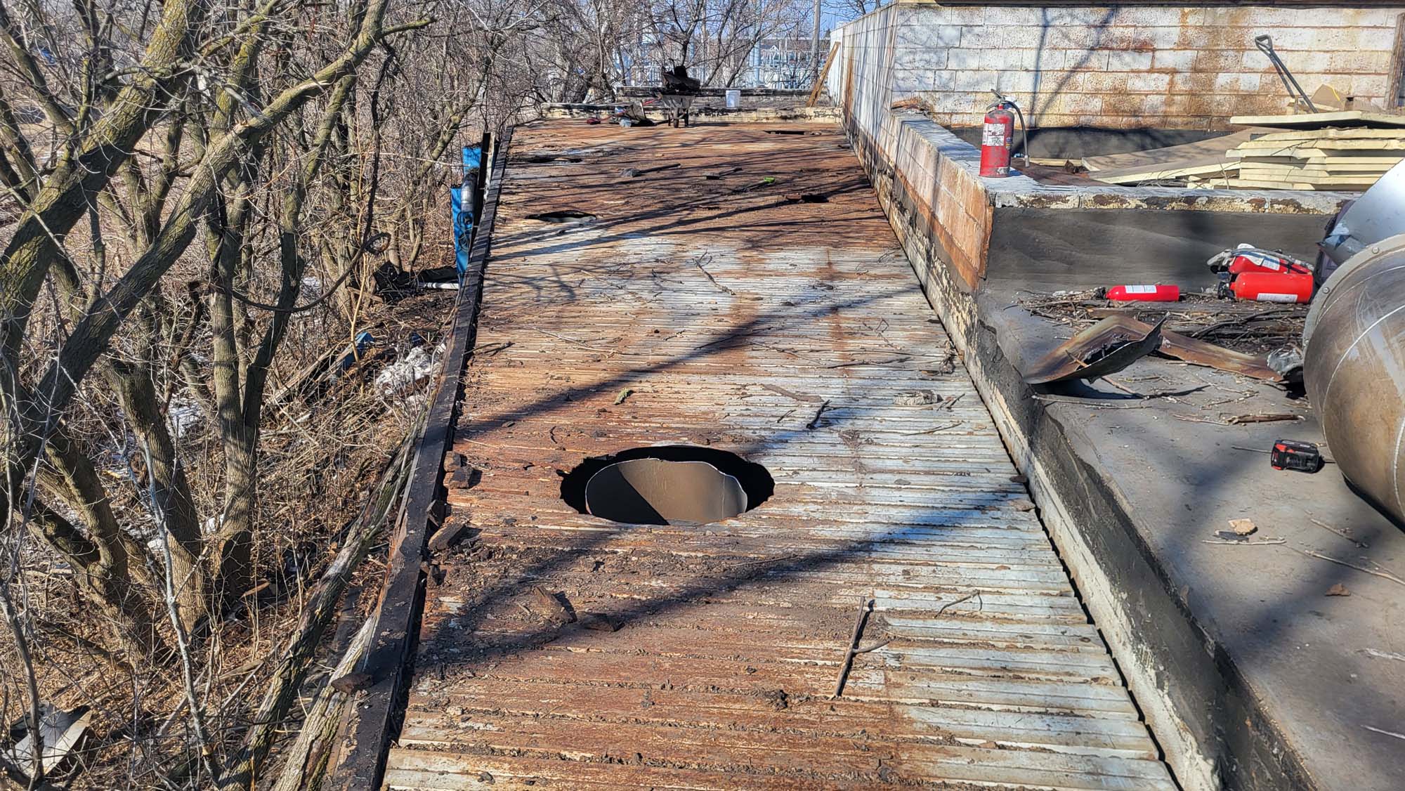 A rusted metal rooftop with a large circular hole in the center, surrounded by debris and scattered tools. Trees are visible to the left, and a fire extinguisher sits on a ledge to the right.