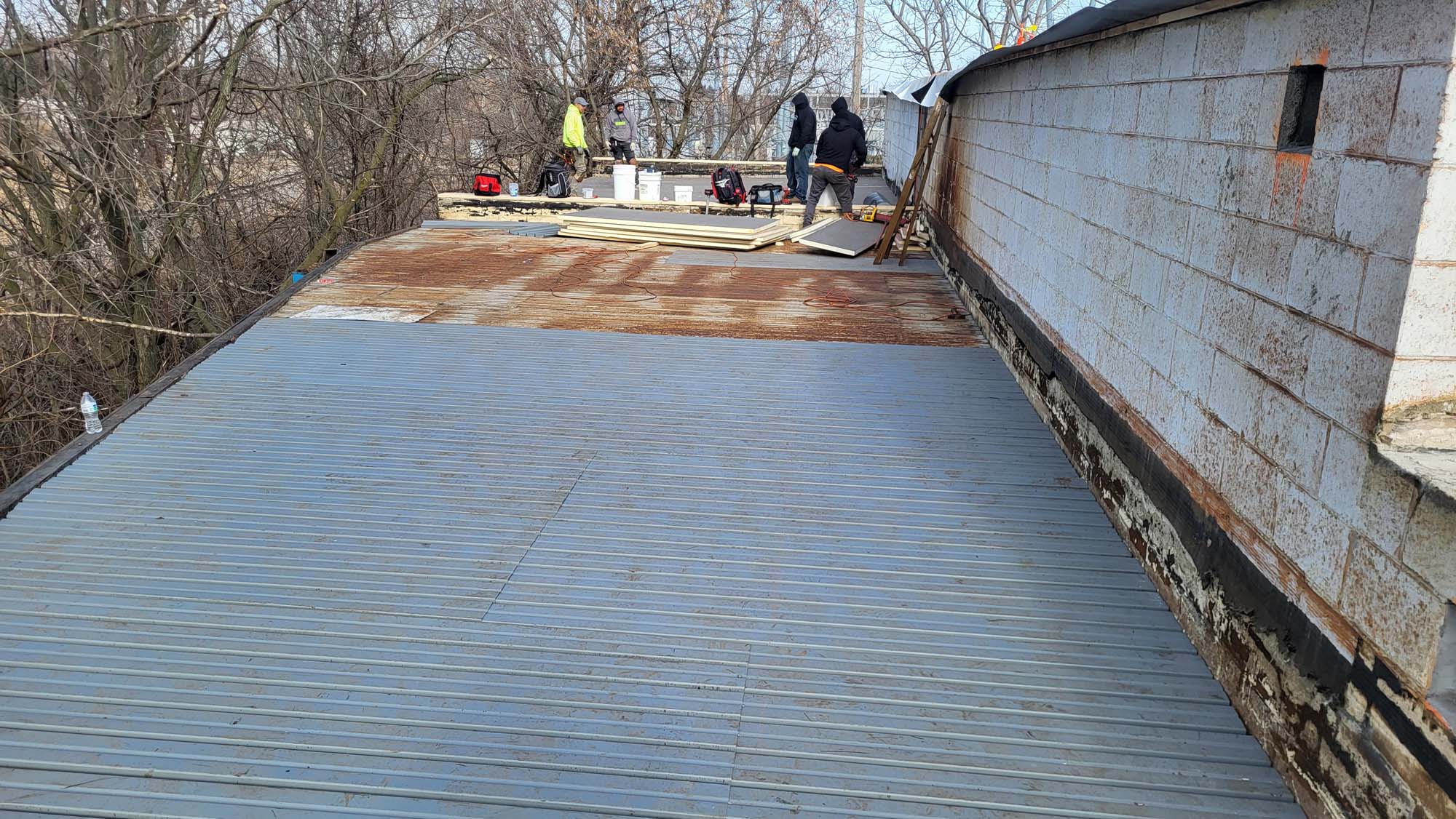 Workers are repairing or installing a metal roof on a flat building. Construction materials and tools are scattered around, and trees without leaves surround the site. The building has a concrete block wall.