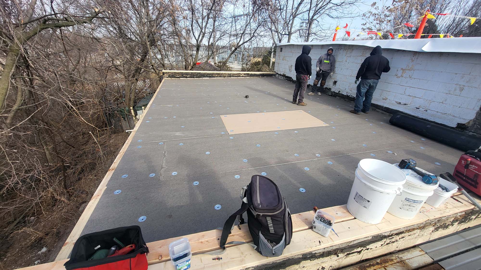 Three people work on a flat rooftop surrounded by trees, attaching roofing materials near a low white wall. Tools, buckets, and bags are scattered on the wooden edge of the roof.