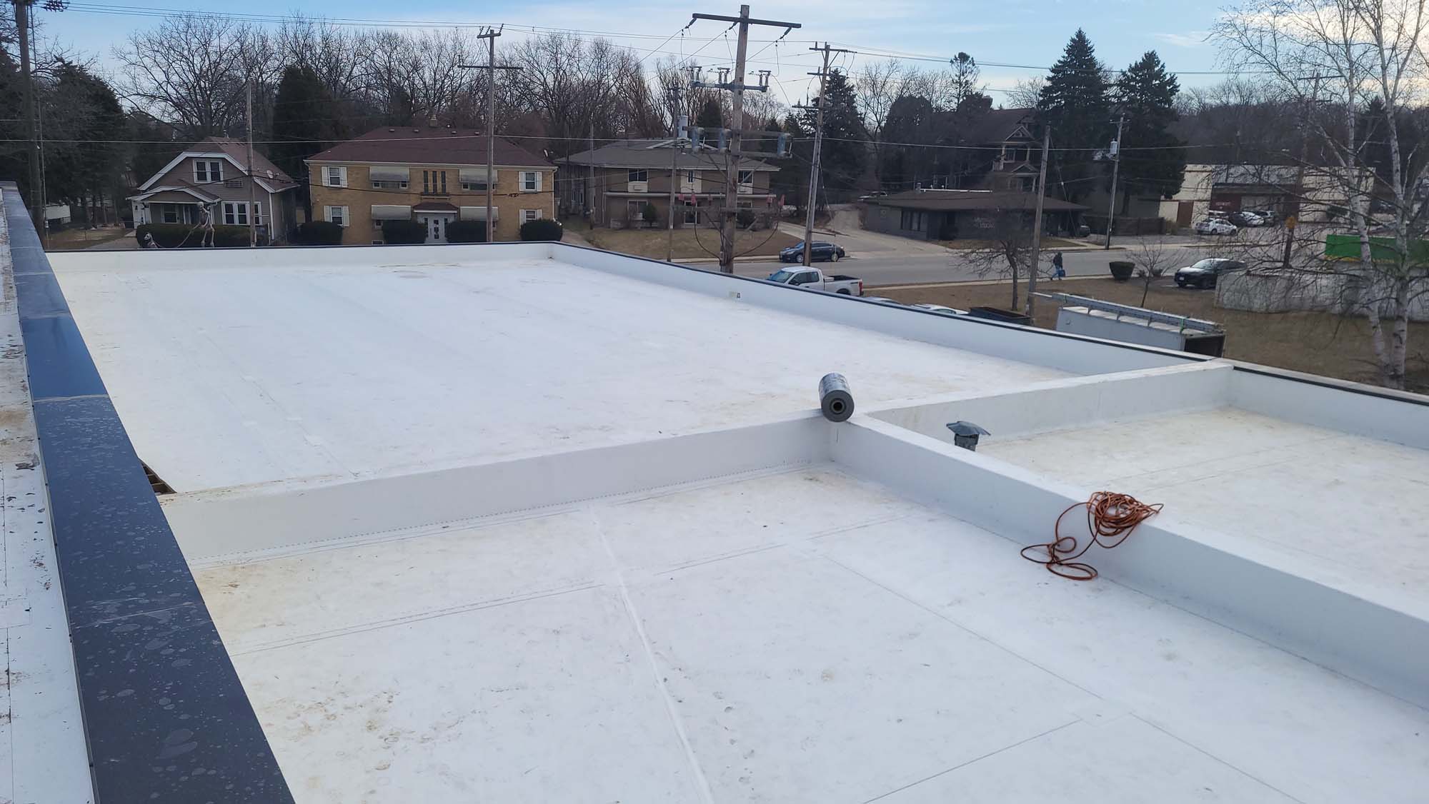 A flat white commercial rooftop with a raised edge, a coiled orange extension cord, a vent pipe, and nearby residential houses and trees in the background.