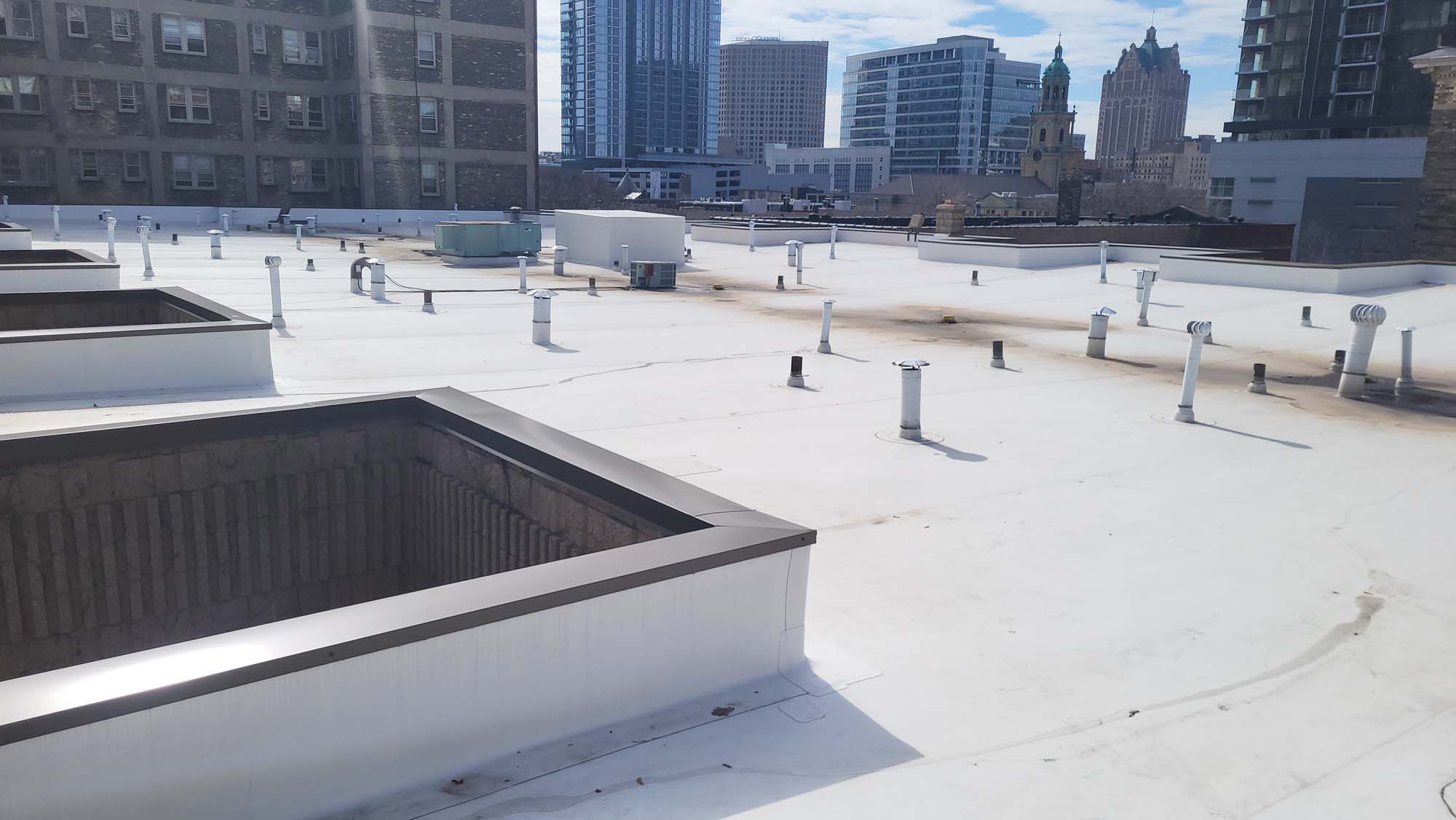 A flat white rooftop with multiple vents, pipes, and HVAC units, surrounded by tall city buildings under a partly cloudy sky.