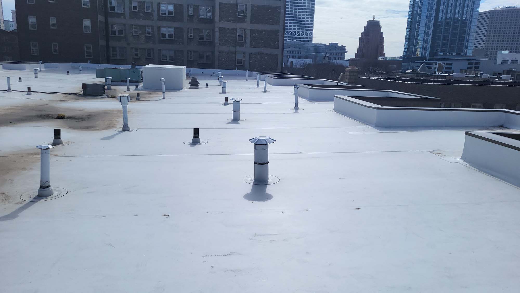 Flat white rooftop with multiple vent pipes and HVAC units, surrounded by tall urban buildings under a partly cloudy sky.