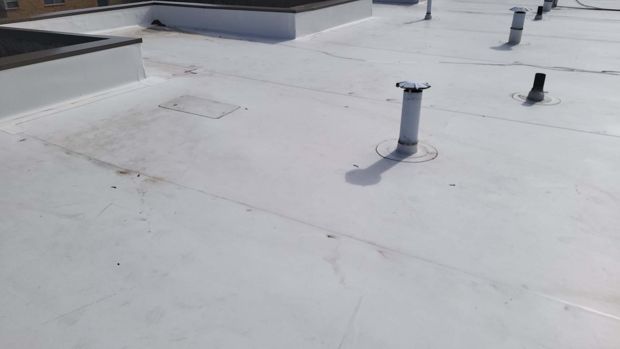 A white flat rooftop with several vent pipes and slight dirt marks, under a clear sky. The surface appears smooth with a few seams and a rectangular hatch visible.
