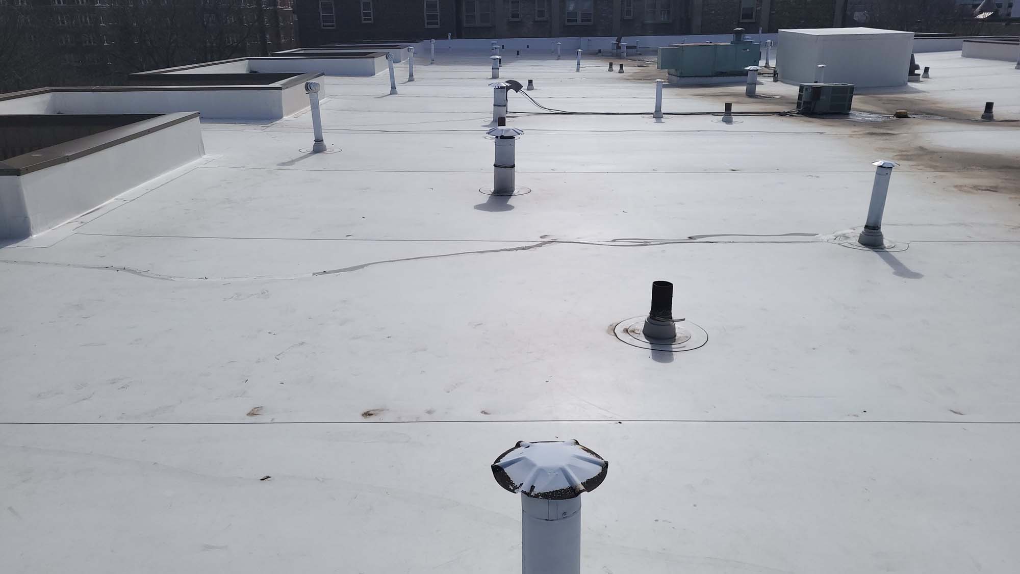 A flat white rooftop with various ventilation pipes, ducts, and HVAC units, under daylight. Some surface cracks and stains are visible across the roof. Trees and buildings appear in the distant background.