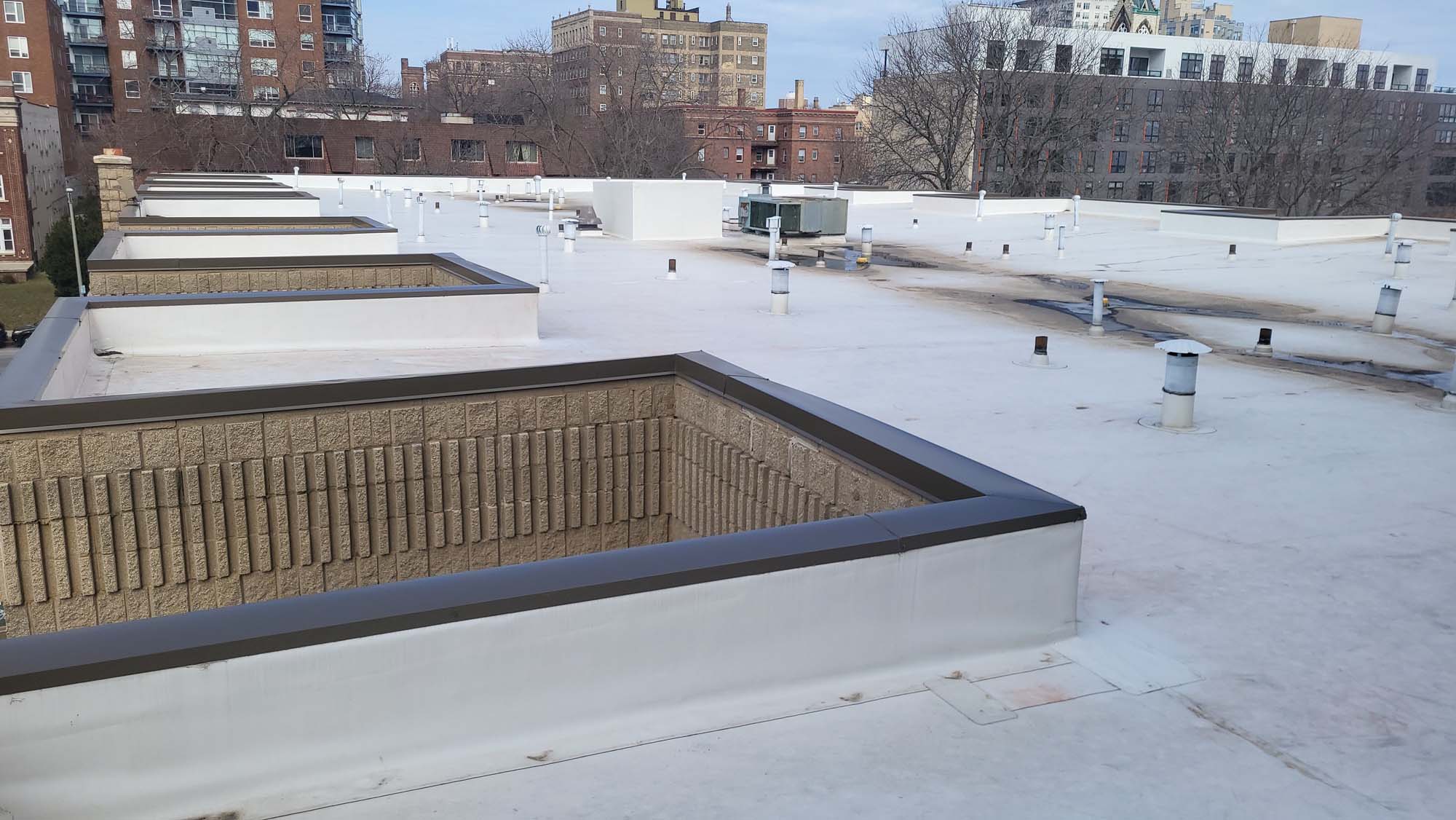 A flat white rooftop with multiple rectangular brick structures and various roof vents, with city buildings and leafless trees visible in the background.