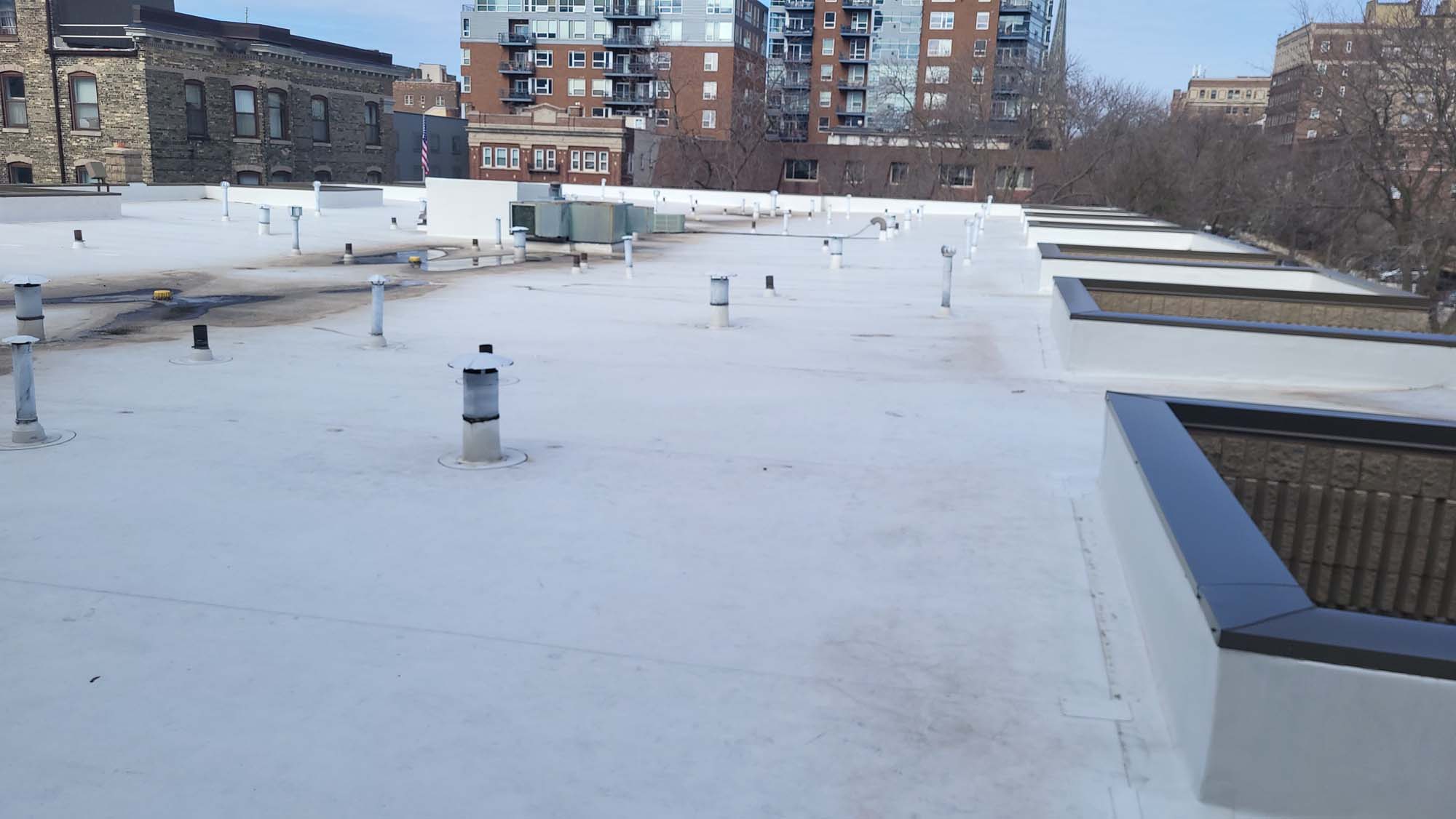 A flat, white rooftop with multiple vents and pipes, surrounded by mid-rise brick and glass buildings on a clear day. Trees without leaves are visible in the background.