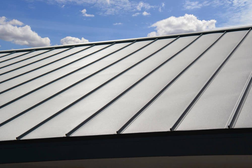 A close-up of a modern metal standing seam roof with raised vertical lines, viewed from an angle against a blue sky with scattered white clouds.