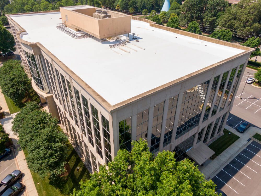 Aerial view of a modern, four-story office building with large glass windows, a flat white roof, and surrounding trees and parking lot on a sunny day.
