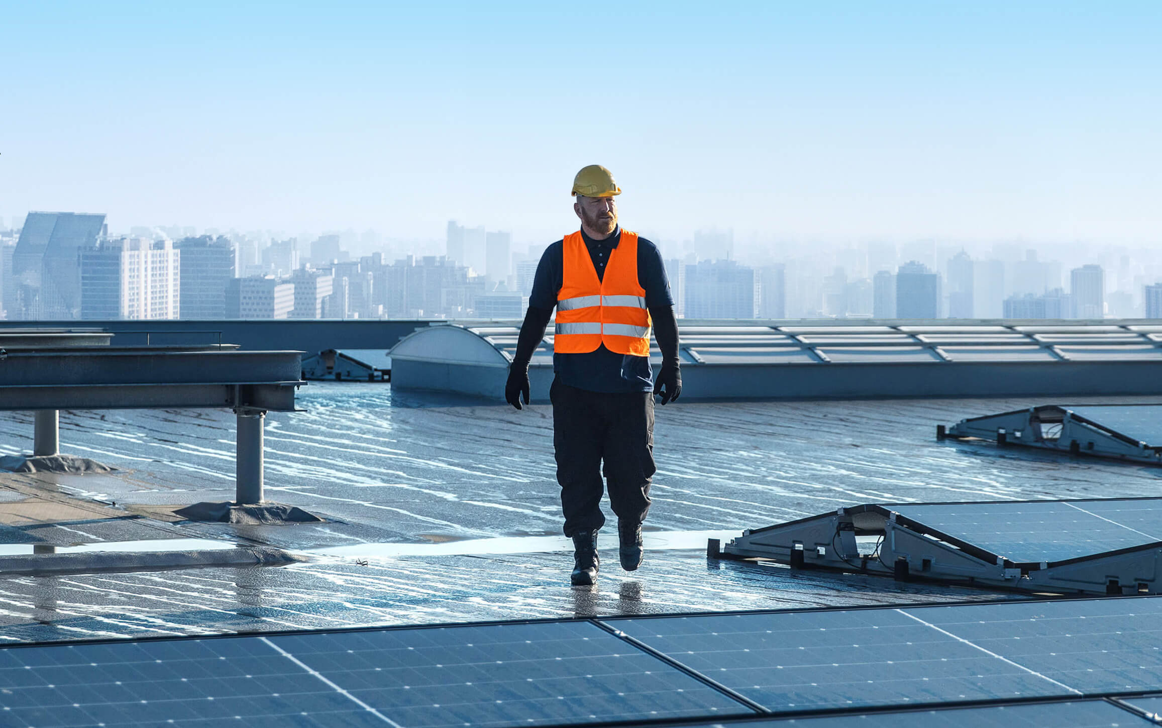 A worker wearing a yellow hard hat and orange safety vest walks on a rooftop covered with solar panels, representing a Brookfield WI roofer specializing in commercial roofing, with a city skyline visible in the background.