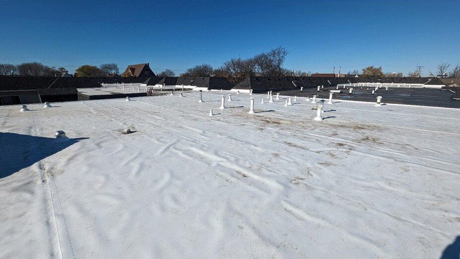 A flat commercial roof covered with white roofing material, featuring multiple vents and pipes. Trees and buildings are visible in the background under a clear blue sky.