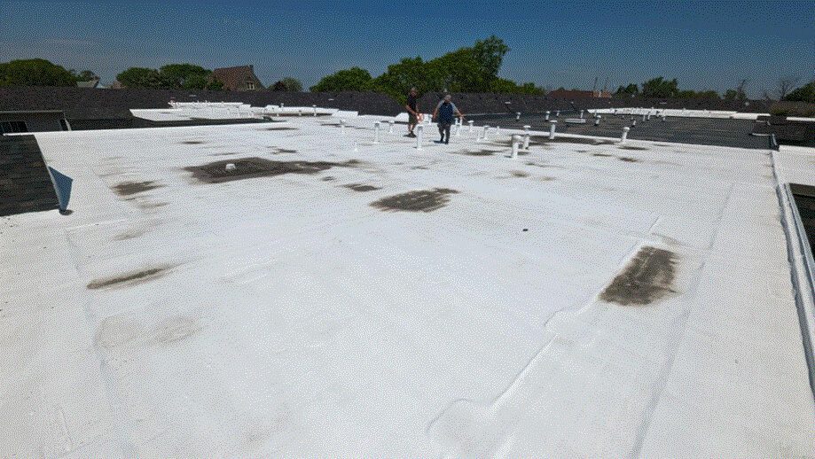 Two people walk on a large, flat, white roof with several pipes and vents; patches of dirt or discoloration are visible on the surface under a clear blue sky.