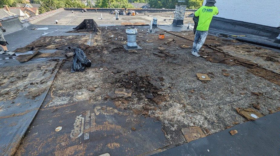 Workers are removing old, damaged roofing material from a flat commercial roof. Debris is scattered around, and one person in a neon shirt is working while another stands nearby. Roofing tools and supplies are visible.
