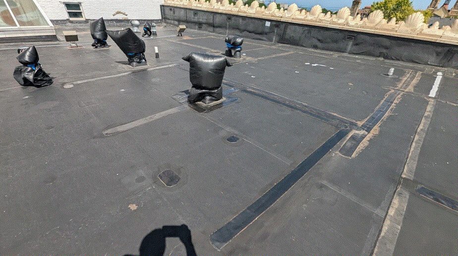 A flat roof covered with black material, featuring several vent pipes wrapped in black plastic. Shadows and patches indicate repairs or maintenance. Decorative stone railing and blue sky are visible in the background.
