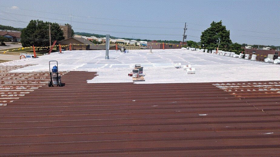 Workers are applying a white coating to the roof of a large industrial building. The front part of the roof is still brown, while the middle and back are mostly covered in white. Equipment and supplies are visible on the roof.
