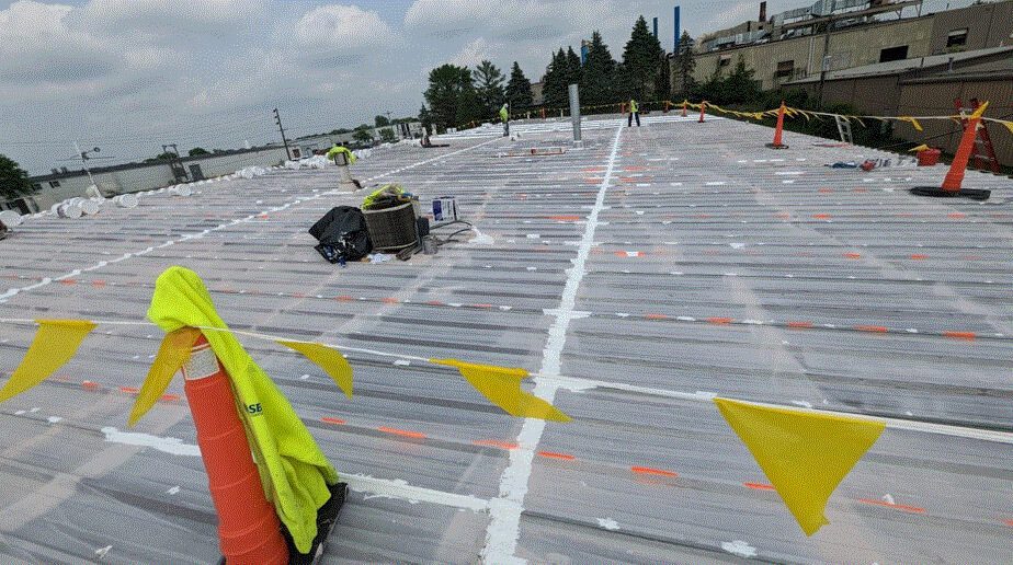 A flat commercial roof under construction is covered with plastic sheeting and white sealant. Yellow safety flags and orange cones mark the perimeter, while workers and equipment are visible in the background.