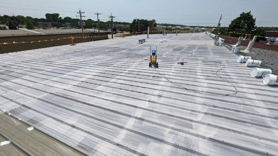 A large industrial metal roof with white coating being applied; equipment, paint rollers, and several buckets are scattered across the roof surface under a clear sky.