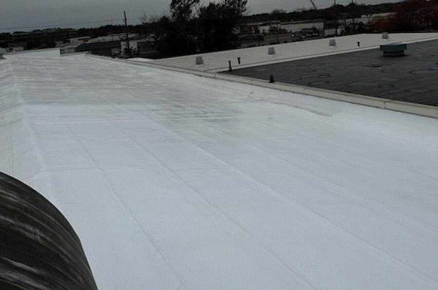 A large flat commercial roof coated with a white reflective material, likely for waterproofing or insulation. The sky is overcast and industrial buildings are visible in the background.