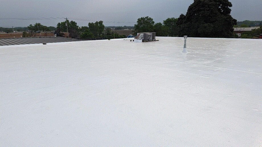 A flat white commercial roof with rooftop equipment and vents, surrounded by trees and buildings under a cloudy sky.