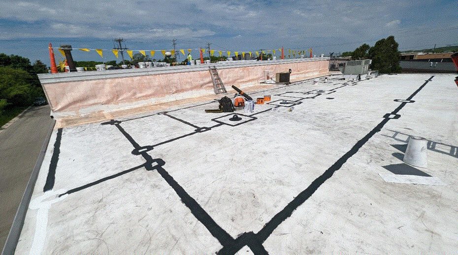 A flat commercial rooftop with black sealant lines, construction tools, a ladder, and safety flags. The sky is partly cloudy and trees are visible in the background.