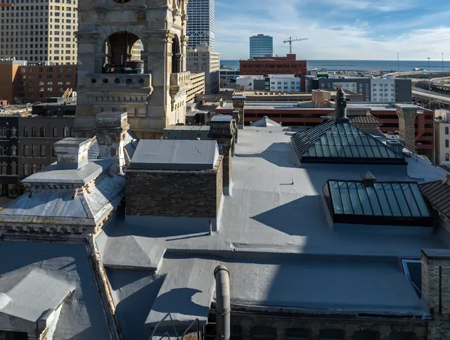 A view from above of historic buildings with sloped rooftops and skylights in an urban city. Modern high-rise buildings and a body of water are visible in the background under a clear blue sky.
