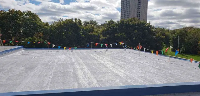 A flat rooftop with a gray surface, surrounded by colorful triangular pennant flags. Trees and a tall building are visible in the background under a partly cloudy sky.