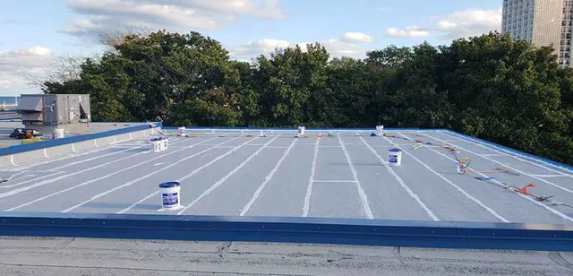 A flat rooftop with buckets of coating material and construction tools spaced around; the roof has white lines marking sections and is surrounded by trees and buildings under a partly cloudy sky.
