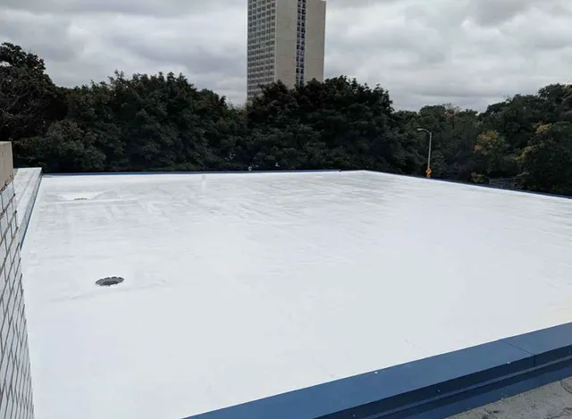 A flat white rooftop with a drain, surrounded by trees and a tall building in the background under a cloudy sky.