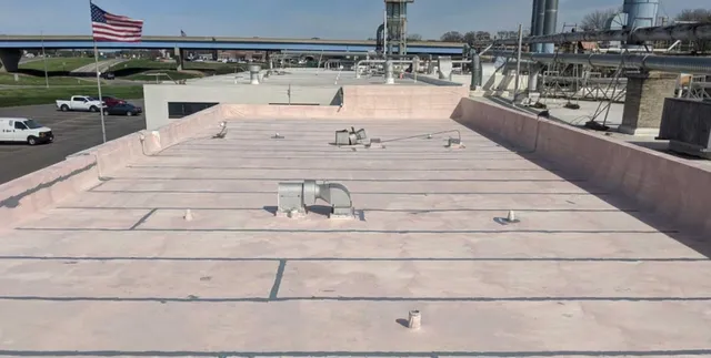 A flat, light-colored industrial rooftop with several vents and pipes, surrounded by low walls. In the background, there are buildings, an American flag, parked vehicles, and an overpass. The sky is clear.