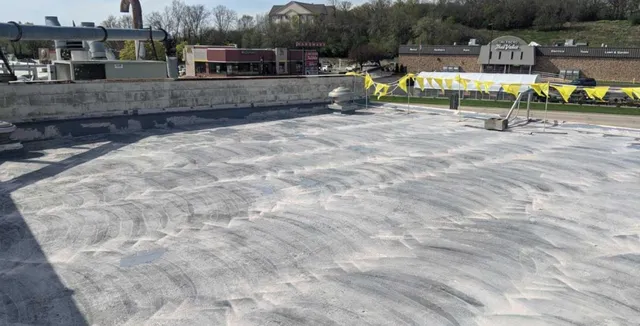 A rooftop with a mostly bare, light-colored surface showing circular cleaning marks. Yellow warning flags line the edge, and HVAC units and surrounding buildings are visible in the background.