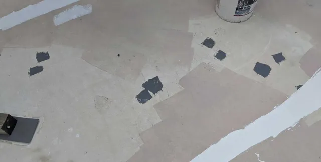 A beige surface with several irregular gray patches, some white paint streaks, and a white bucket in the upper right corner. The area appears to be undergoing repair or renovation.