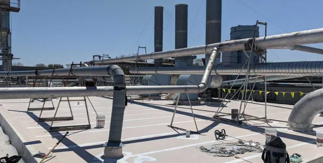 Pipes and ducts run across an industrial rooftop with equipment and ventilation towers in the background. Buckets, hoses, and construction tools are scattered across the flat roof under a clear sky.