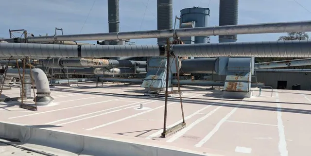 A flat industrial rooftop with large metal ventilation ducts, pipes, and exhaust stacks, all exposed under a clear sky. The roof surface is light-colored and marked with white lines.