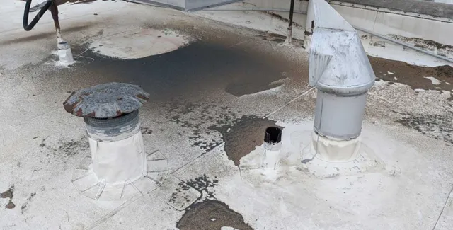 A flat rooftop with vents and dark stains surrounding them, suggesting possible water or chemical leakage. The surface is worn and weathered, with patches of discoloration and sealant around the base of the vents.