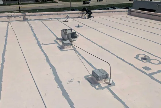 A person kneels on a flat, light-colored rooftop near some vents and equipment, with electrical and piping lines visible. Streets, grass, and parked cars are in the background.