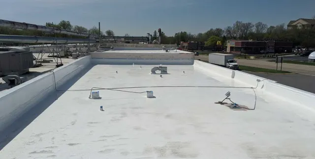 A flat, white commercial rooftop with various vents, pipes, and equipment, surrounded by low walls. Buildings, trees, and a road are visible in the background under a clear blue sky.