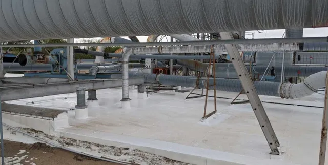 Large metal pipes and ducts run across an industrial rooftop with a white surface. Some pipes show signs of ice or frost, and metal supports hold up several pipes over the roof. The sky is cloudy.