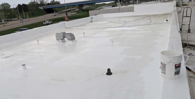 A flat white commercial roof with vents, utility structures, and a white bucket on the right side. Nearby roads, a highway overpass, and grassy areas are visible in the background.