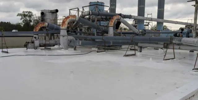 Large industrial pipes and ducts run across a flat, white rooftop on a cloudy day. Some pipes are rusted, and there are metal railings and industrial structures visible in the background.
