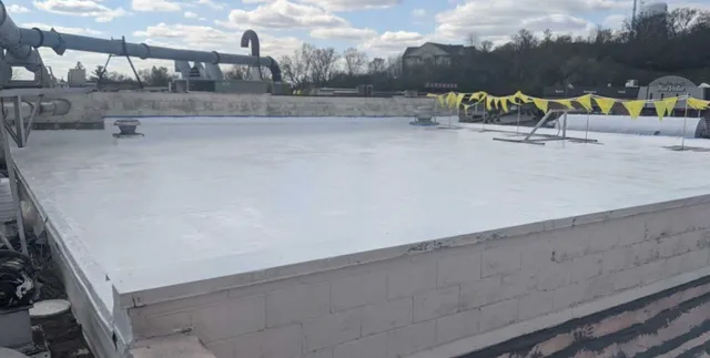 A flat white rooftop with pipes and vents, surrounded by yellow caution tape and buildings visible in the background under a partly cloudy sky.