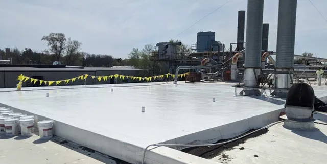 A flat industrial rooftop with white coating, surrounded by yellow caution flags. Several vents and pipes are visible, along with buckets near the edge, and industrial structures appear in the background under a clear sky.