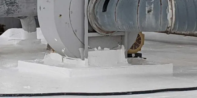 A close-up view of industrial ventilation equipment mounted on a flat, white rooftop, featuring a metal duct, a fan or blower unit, and some patched or sealed areas around the base.