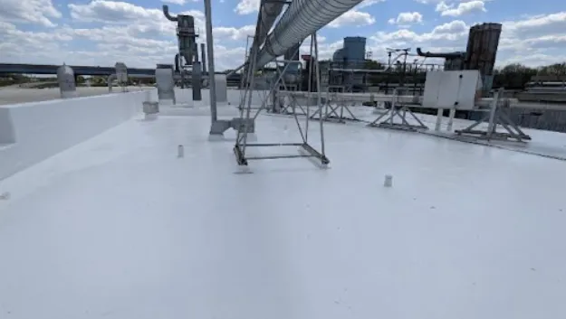 A flat white rooftop with industrial equipment, pipes, and vents under a partly cloudy sky, with buildings and structures visible in the background.