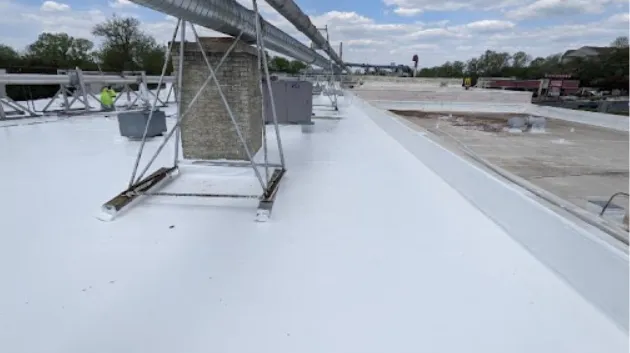 A flat commercial rooftop with a white reflective coating, various HVAC units, vents, and a few workers visible in the background under a partly cloudy sky.
