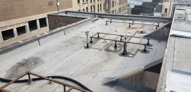 A flat rooftop with several metal pipes and railings, some rusted, scattered water stains, and nearby brick buildings in an urban setting under daylight.