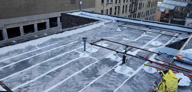Flat urban rooftop with white waterproof sealant applied in lines and patches, several pipes and vents protruding, metal scaffolding, and a yellow construction bag; surrounding buildings visible in the background.