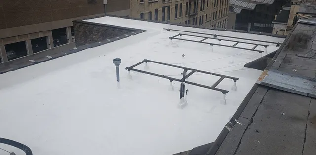 A flat white rooftop with various metal pipes, vents, and structures, surrounded by taller buildings in an urban setting. The roof appears recently coated or covered, and rooftops of other buildings are visible in the background.