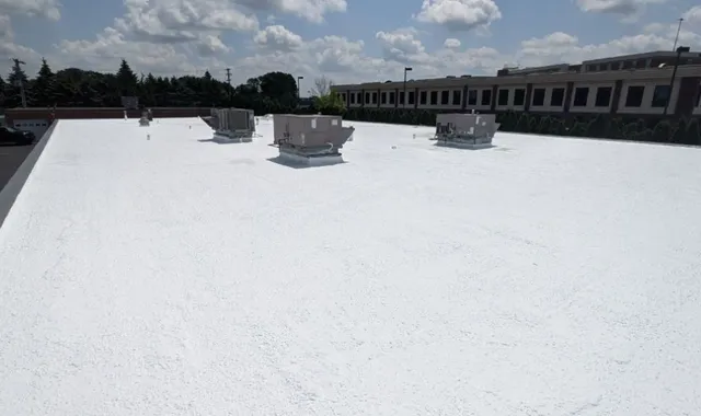 A large, flat white rooftop with several HVAC units under a partly cloudy sky, next to a building with rows of windows and surrounded by trees.