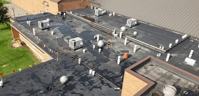 A flat, black-roofed building with numerous white vents, HVAC units, and pipes. The roof shows patches of water and scattered maintenance equipment. A green lawn and brown brick walls are visible along the edges.